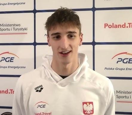 Ksawery Masiuk, polski pływak specjalizujący się w stylu grzbietowym, w dynamicznym ujęciu podczas wyścigu, odbijający się od ściany lub wykonujący nawrót, z rozpryskującą się wokół wodą.
