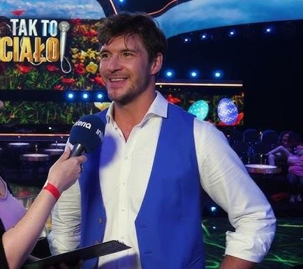 Mikołaj Roznerski uśmiecha się, trzymając kubek kawy, w swobodnej interakcji z fanami, w tle widać kawiarnię.