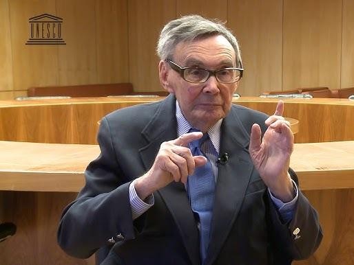 Marian Turski przemawiający podczas uroczystości upamiętniającej Holokaust, z symbolicznym tłem obozu Auschwitz-Birkenau lub Muzeum POLIN.