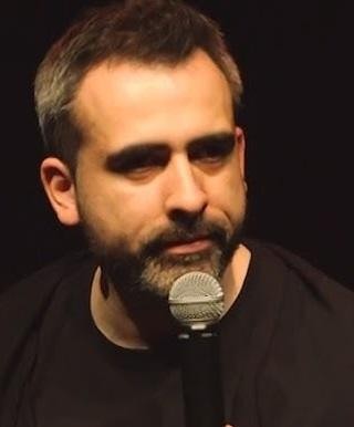 Abelard Giza podczas występu stand-upowego, z mikrofonem, w energicznej pozie, na tle rozmytej publiczności i świateł sceny.