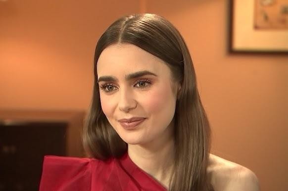 Lily Collins uśmiechająca się subtelnie, ubrana swobodnie, ale stylowo, w ciepłym, domowym otoczeniu.