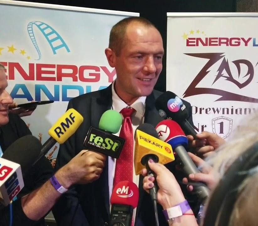 Marek Goczał w stroju rajdowym, stojący obok pojazdu SSV, z tłem łączącym park rozrywki Energylandia i tor rajdowy.