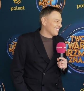 Krzysztof Ibisz w studiu telewizyjnym, z mikrofonem, w tle elementy scenografii programu rozrywkowego, uśmiechnięty i pełen energii.
